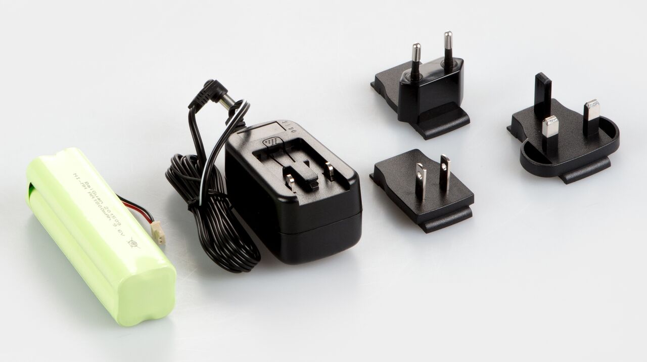 Akkubetrieb Set zum einbauen in die Waage auf weißem Hintergrund. Das Set besteht aus einer Batterie in hellgrünem Kunststoff eingeschweißt, einem Stecker mit aufgefaltetem Kabel und drei Adapter für EU, UK und US.