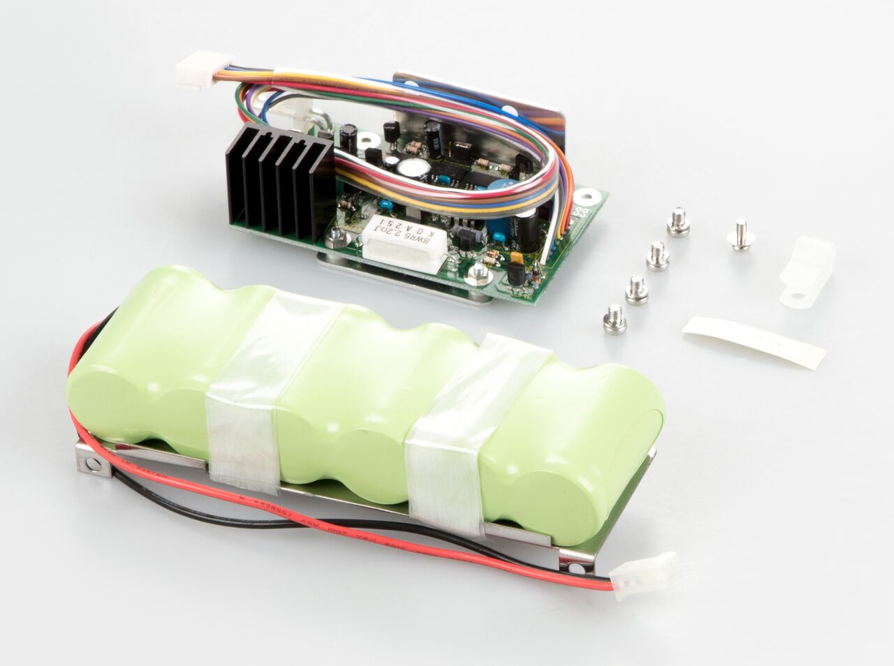 Akkubetrieb Set zum Einbau in die Waage auf weißem Hintergrund. Das set besteht aus in hellgrünem Kunststoff eingeschweißten Batterien, eine Leitplatte mit einem bunten Kabel und mehrere Schrauben.