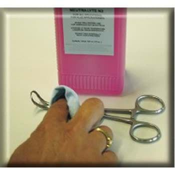 Eine Hand reinigt mit einem Tuch eine silberne Zange. Dahinter steht eine pinke Flasche mit weißem Etikett. Beide liegen auf eine gelbliche Oberfläche.