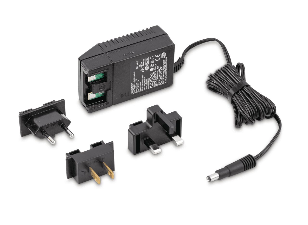 Vier schwarze Stecker auf weißem Hintergrund. Das große Netzteil liegt auf der Seite, das Kabel zusammengebunden daneben. Davor liegen drei Adapter: einen für Europa, einen für die USA und einen für Großbritannien.